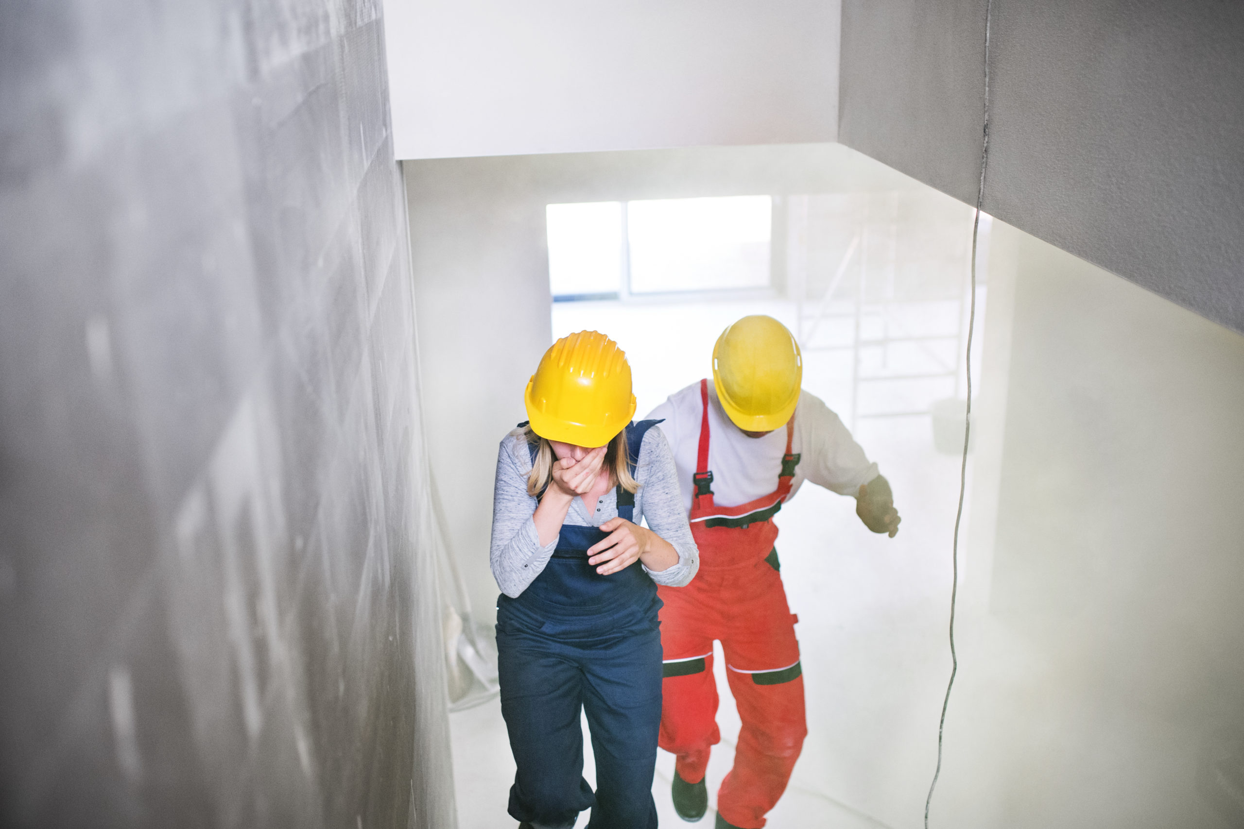 A woman and a man in construction clothes are running upstairs through dust while coughing at a worksite.